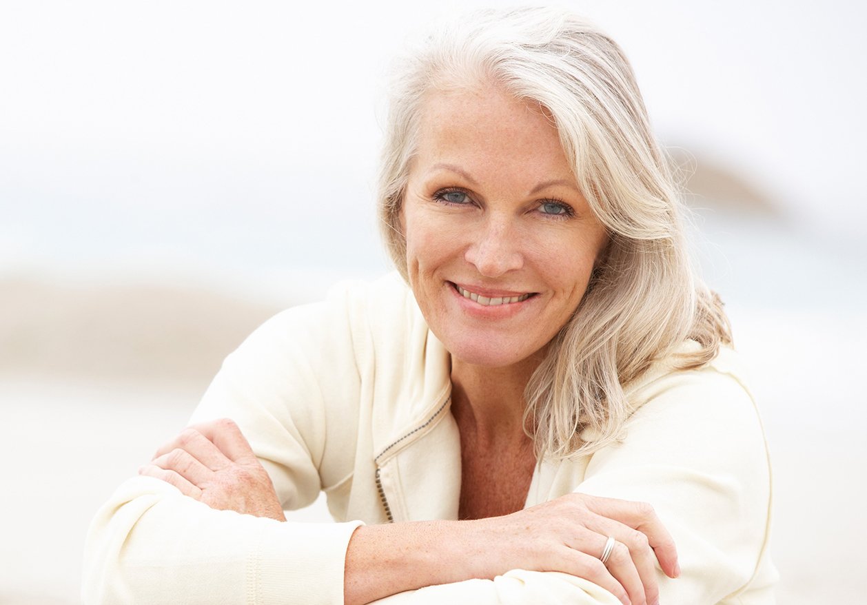 An older woman with gray hair smiles at the camera while sitting on the beach, showing her successful treatment of menopause symptoms.