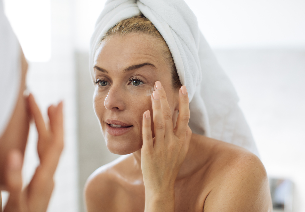 A woman with a towel in her hair applies skincare to her face while looking in the mirror.
