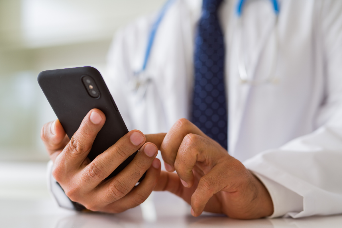 A doctor in a lab coat with a stethoscope touches the screen of his smart phone to indicate telemedicine.