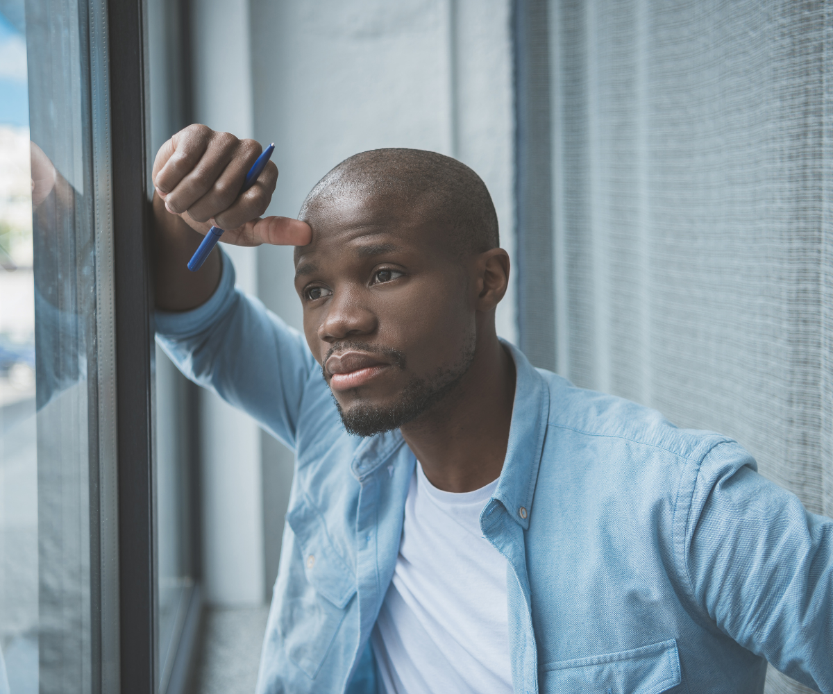 A Black man stares pensively out of the window, considering treatments for erectile dysfunction.