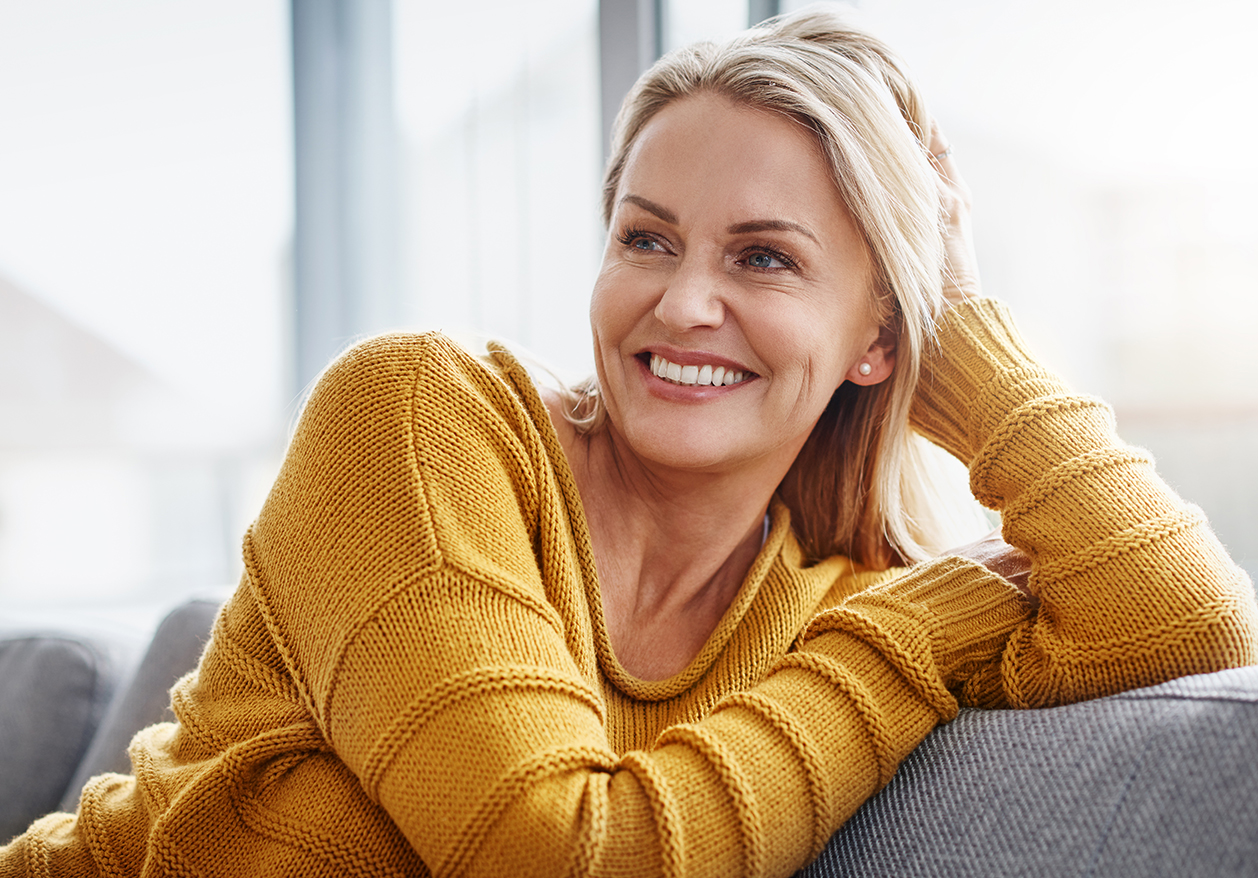 A mature woman sits on a couch smiling in a yellow sweater, showing the success of her hormone therapy.