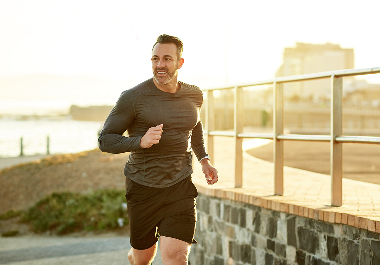 A man smiles as he runs near a bridge and body of water, showing the benefits of Testosterone Replacement Therapy online.