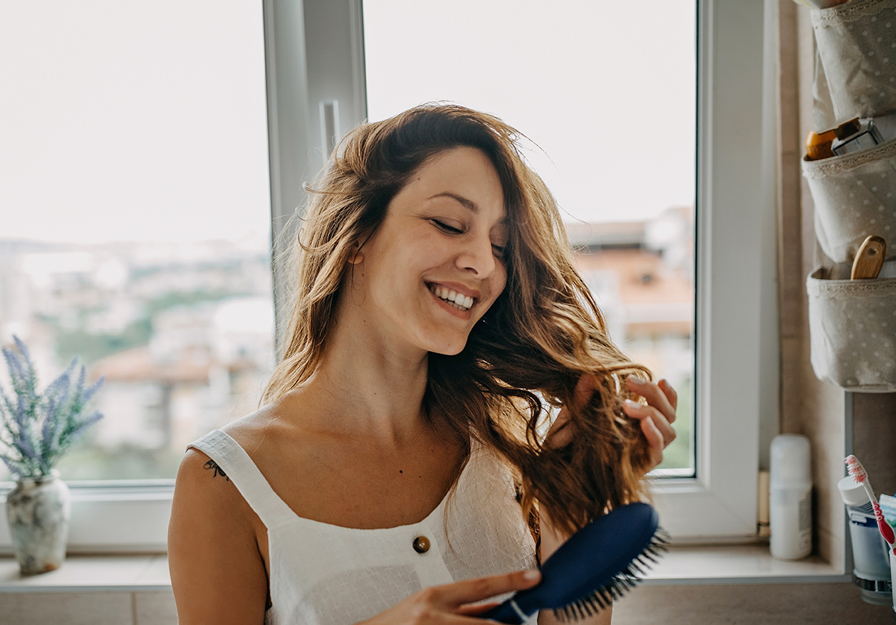 A woman with brown wavy hair brushes her hair while smiling because her hair loss treatment is working.