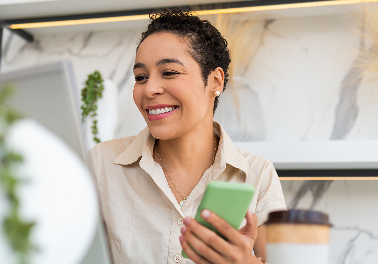 A black woman holds a phone and looks at her laptop while smiling to book telemedicine primary care online.