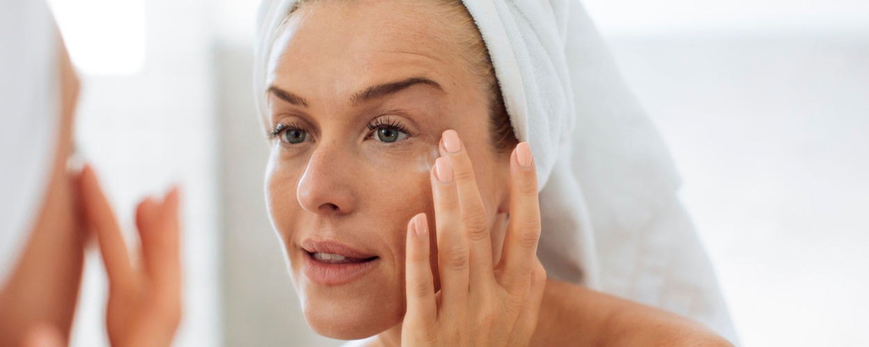 A woman with her hair wrapped in a bath towel looks at the benefits of her skincare in the mirror.