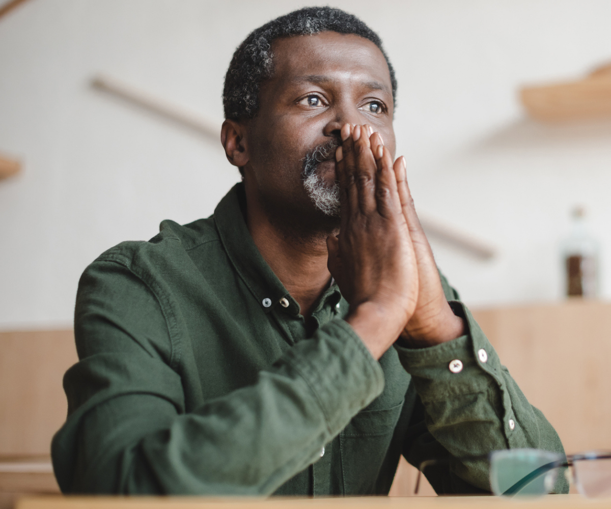 A man stares pensively out of the window with his hands in front of his mouth.