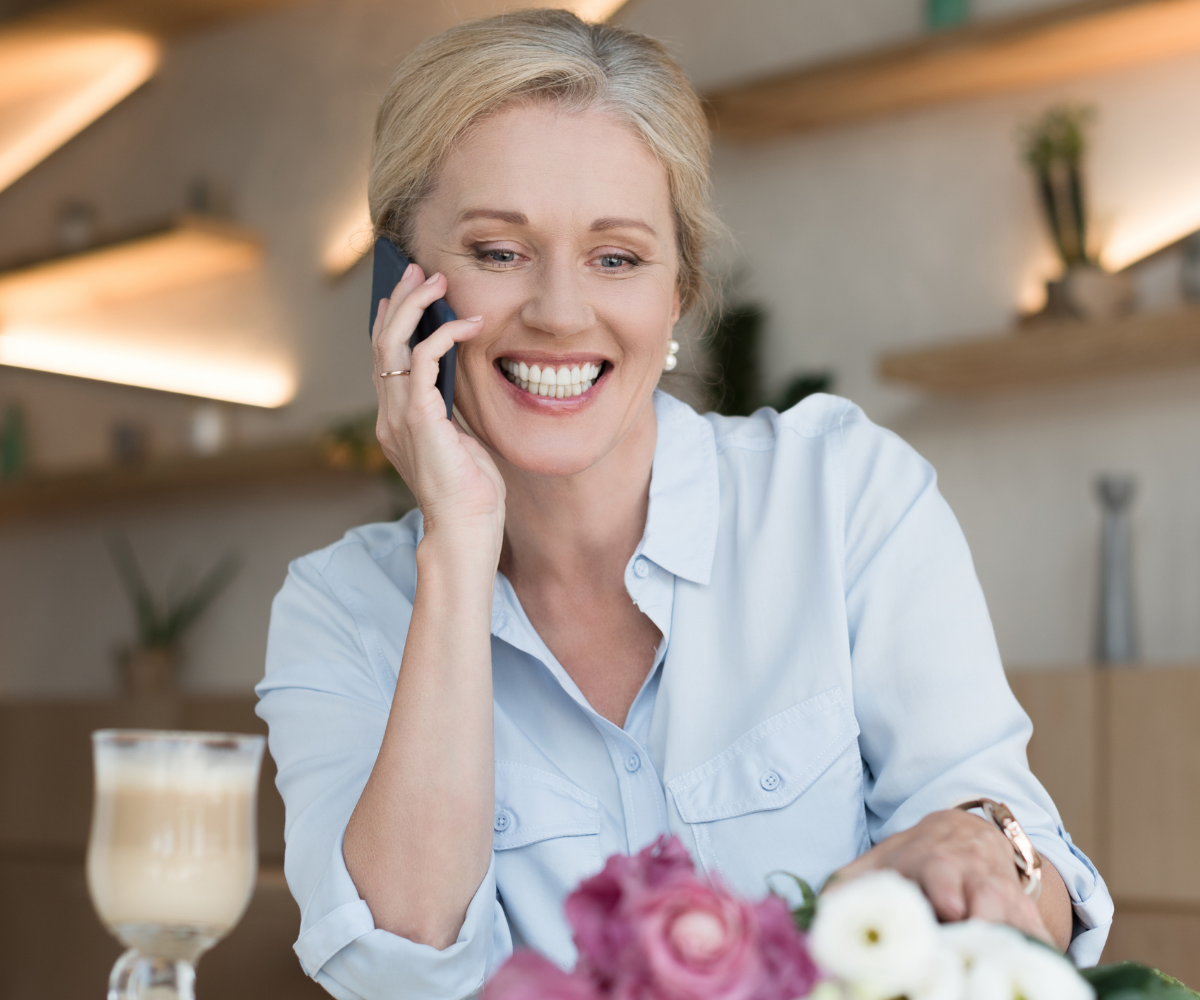 A woman of menopause age smiles as she talks on the phone with a coffee and flowers on the table in front of her.