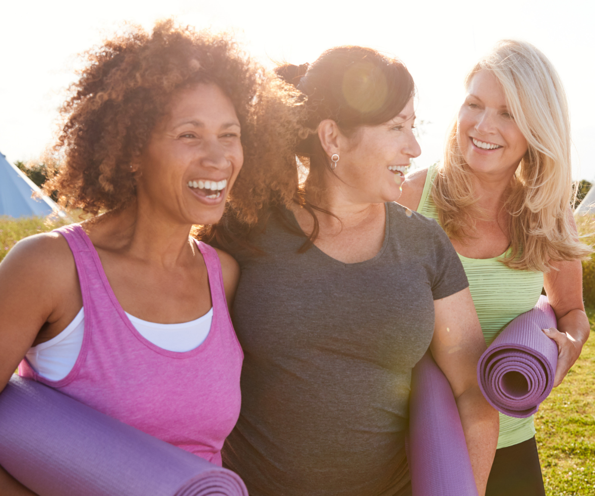 A group of women of menopause age in athletic gear smile as they carry yoga mats.