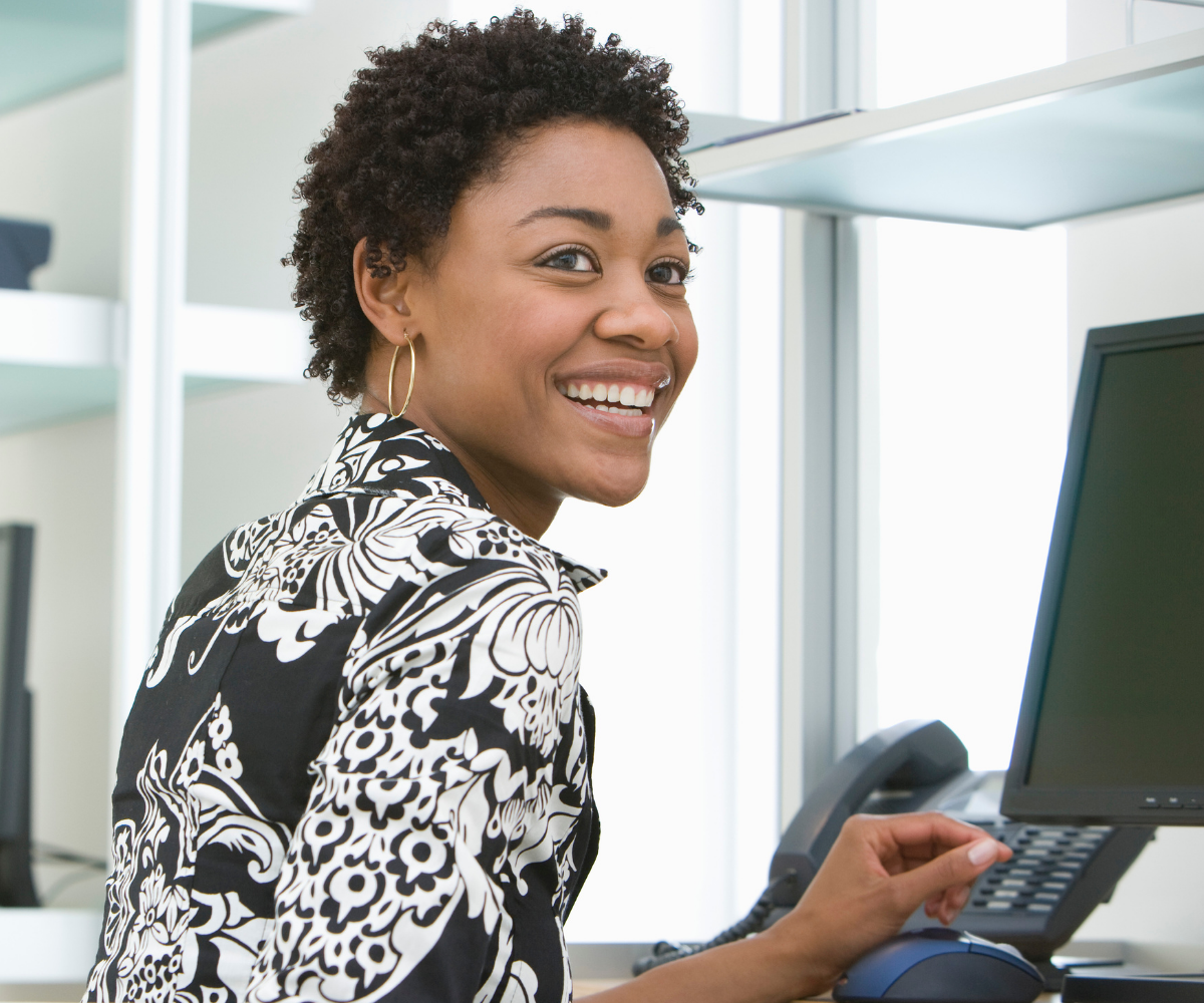 A woman of menopause age sits in front of a computer and office phone and smiles as she looks back from her work desk.