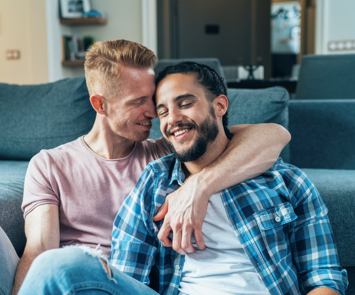 Two gay men embrace while sitting in front of the couch.