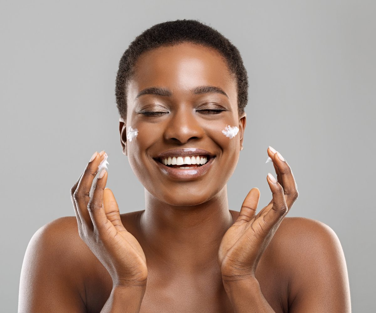 A black woman smiles as she applies skincare to her cheeks.