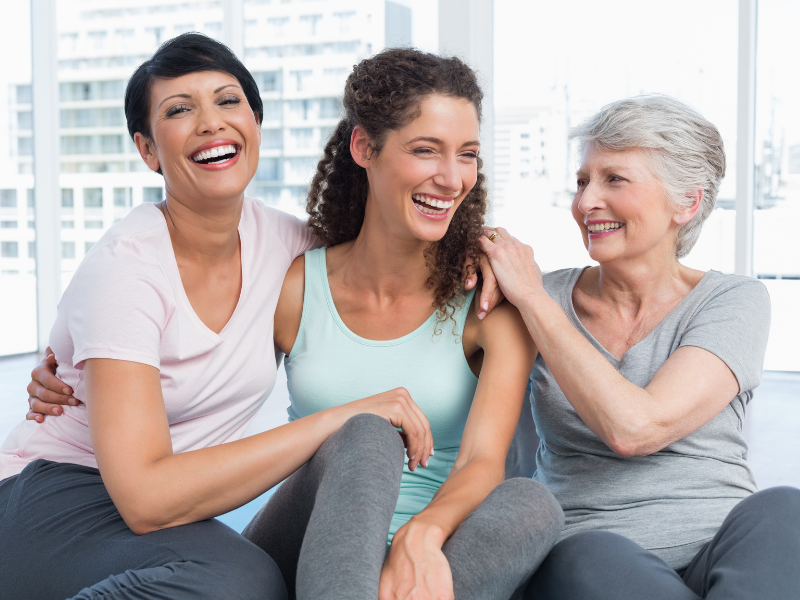 Three woman of various ages sit on a couch and laugh together, showing the benefit of hormone therapy for women.