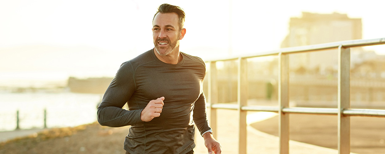 A man smiles as he runs near a bridge and body of water, showing the benefits of Testosterone Replacement Therapy online.