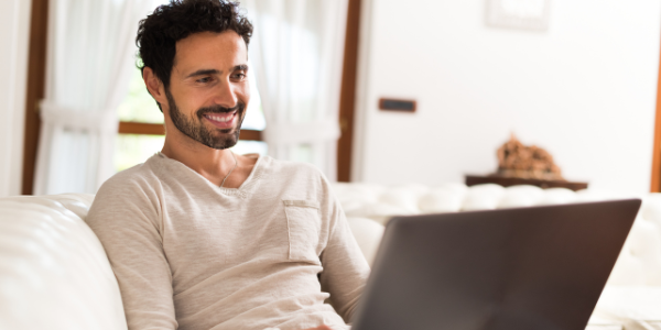 A man with dark hair and a beard smiles as he looks at his laptop.