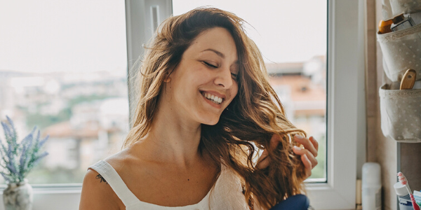 A woman with brown wavy hair brushes her hair while smiling because her hair loss treatment is working.